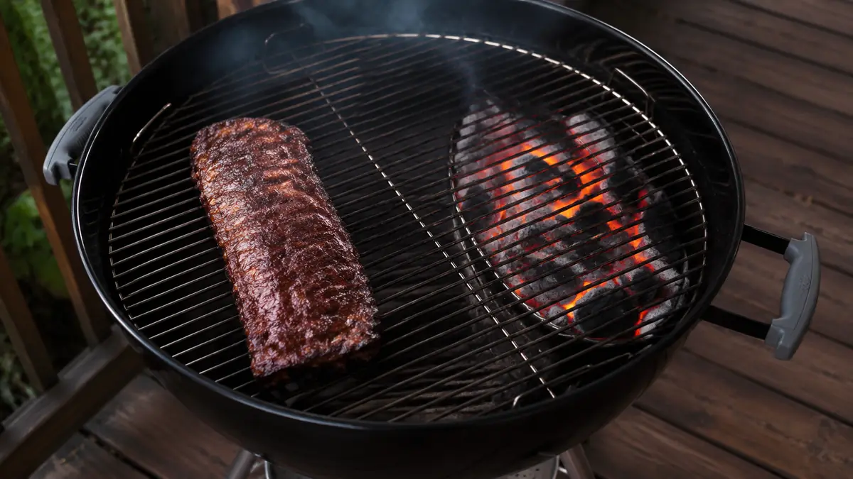 A charcoal kettle grill set up with coals banked on one side and ribs resting on the indirect side