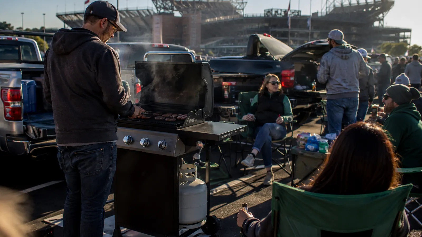 Coleman RoadTrip 285 portable propane grill set up in a stadium parking lot with tailgaters and trucks in the background