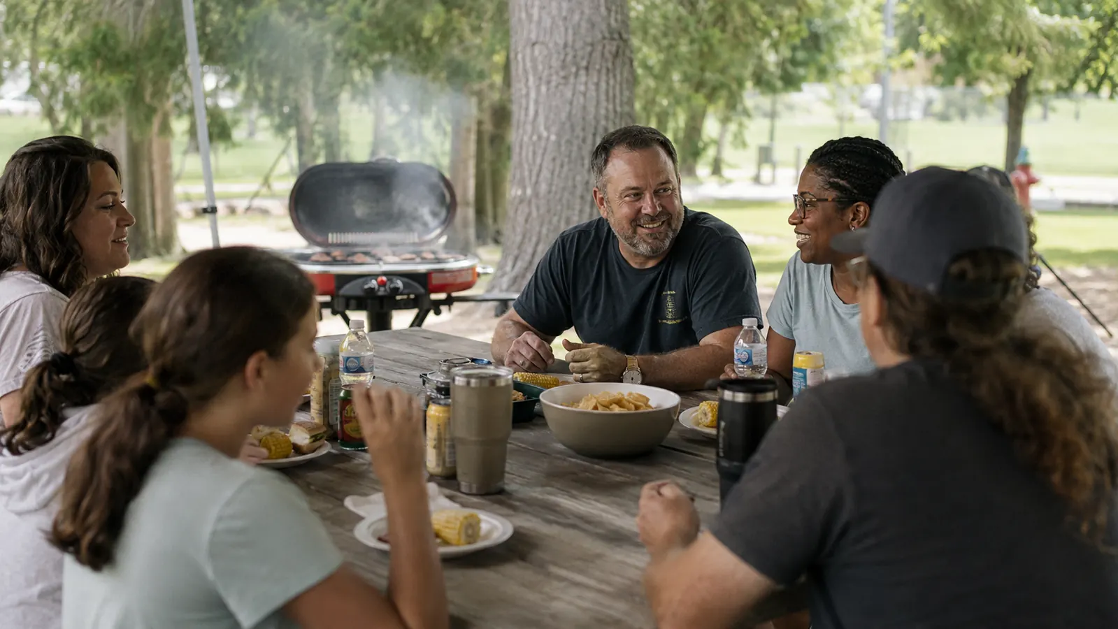 Six people gathered around a picnic pavilion table eating burgers and corn on the cob with the Coleman grill visible nearby