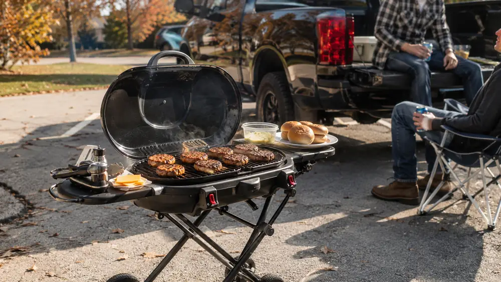 A Coleman RoadTrip 285 propane grill set up on its wheeled stand at a gravel tailgate lot with a truck parked behind it